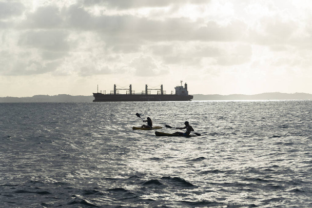 Salvador, Bahia, Brazylia - 21 marca 2021: Ludzie w dwóch kajakach pływający w zatoce wszystkich świętych obok dużego statku. - Zdjęcie, obraz