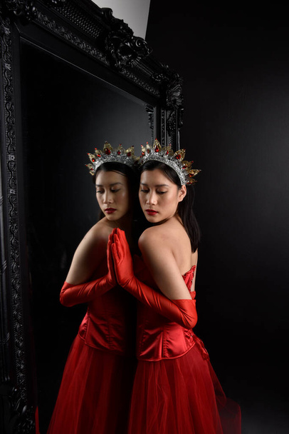 Full length  portrait of beautiful young asian woman wearing red corset, long opera gloves and ornate crown headdress. Graceful posing against a full length mirror with a dark studio background. - Foto, afbeelding