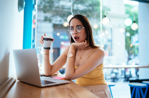Amazed female user with coffee to go watching online webinar video via app on netbook technology, wonder hipster girl in eyewear reading publication in social media networks feeling shocked with news - Фото, изображение