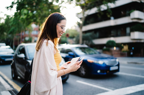 Happy female tourist checking received mobile notification smiling at urban street, cheerful hipster girl reading positive web publication during social networking via smartphone technology - 写真・画像