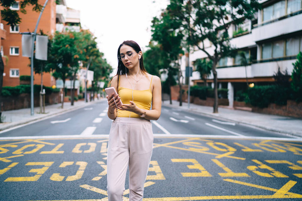 Caucasian hipster girl in optical glasses and headphones tracking travel gps via smartphone application, young female user listening music and networking social media at urban road in city downtown - Фото, зображення