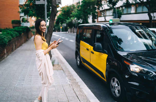 Fröhliches Hipster-Mädchen in Brille mit Handy-Gadget für Online-Buchung Taxis auf der Suche nach Transport in der Nähe der Stadtstraße mit Verkehr, glückliche Millennial Generation Z lächelt im Freien - Foto, Bild