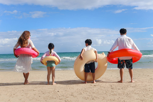 Dzieci z rodzicem noszącym kolorowy nadmuchiwany pierścień pływacki biegnący do błękitnego morza, dzieci i dorośli w kolejce od tyłu na tropikalnej piaszczystej plaży, rodzina spędzająca razem czas na wakacjach.  - Zdjęcie, obraz