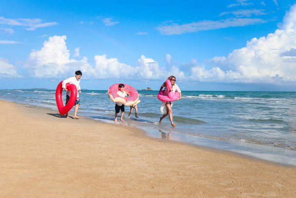 Kinder mit Eltern tragen bunte aufblasbare Schwimmringe, die am Sommerstrand laufen, Kinder und Erwachsene am tropischen Sandstrand, glückliche Familien verbringen Zeit und Spaß zusammen im Sommerurlaub.  - Foto, Bild
