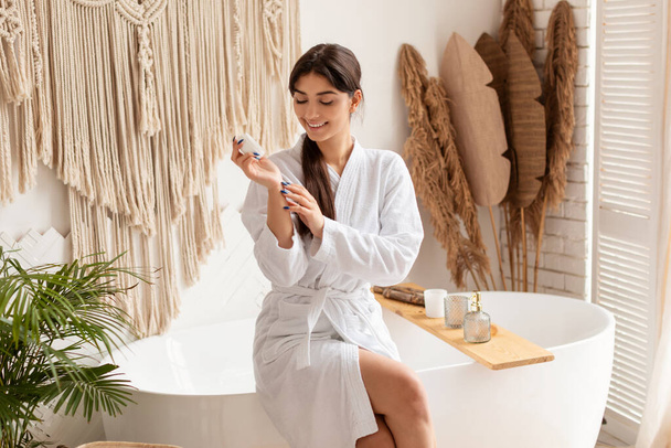 Ευτυχισμένη γυναίκα εφαρμογή κρέμα χεριών κάθεται στο σύγχρονο μπάνιο - Φωτογραφία, εικόνα