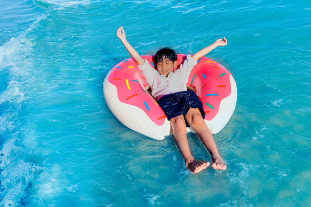 Šťastný chlapec s barevným nafukovacím plaveckým kroužkem plovoucím na hladině a vlnou tropického modrého moře, který tráví čas s dětmi a baví se na letních prázdninách, cítí se dobře a relaxuje o víkendu.  - Fotografie, Obrázek