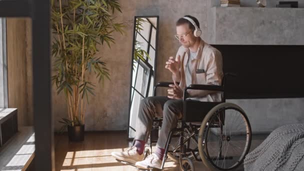Ein glücklicher kaukasischer Mann sitzt im Rollstuhl in einer Wohnung, trägt Über-Ohr-Kopfhörer, hört Musik und imitiert das Spielen auf dem Saxophon - Filmmaterial, Video