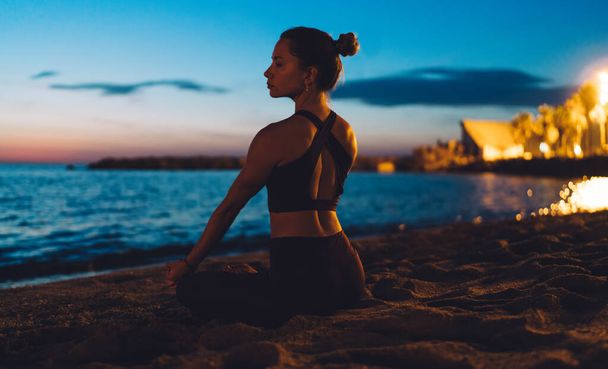 Caucasian female in tracksuit sitting in asana for seeking mindfulness and enlightenment during practice retreat, calm woman enjoying relaxation and vitality during yoga training at seashore coastline - Photo, Image