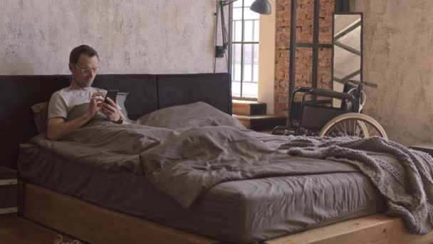 Широкий кавказский мужчина лежит в постели, используя смартфон, инвалидное кресло стоит на заднем плане в дневное время - Кадры, видео
