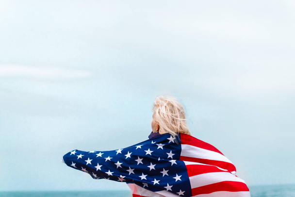 Mujer rubia caucásica sosteniendo bandera americana apoyada en la espalda y mirando al mar. Bandera de EE.UU. para el día de la independencia - Foto, imagen