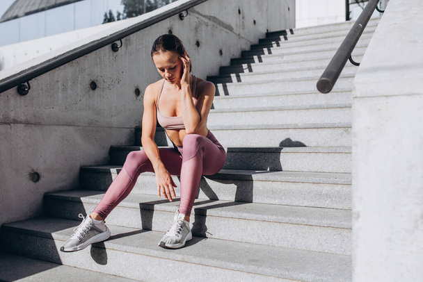 Junge schlanke sportliche Kaukasierin in Sportkleidung ruht sich nach dem Training auf der Straße aus und sitzt auf der Treppe - Foto, Bild