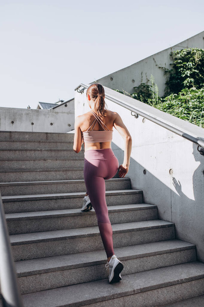 Back view. Young slim sportive caucasian woman working out, doing fitness, yoga on the stairs on city street. - Foto, afbeelding