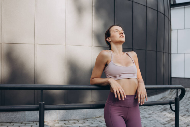 Young slim sportive caucasian woman in sportswear with closed eyes resting after training on the city street. - Photo, Image