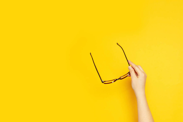 Person hand holds glasses for vision on a yellow background with copyspace, top view, flat lay - 写真・画像