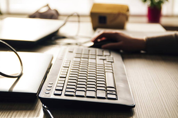Um retrato de um teclado de computador deitado em uma mesa de madeira com alguém usando um mouse no fundo embaçado, a pessoa está usando o PC em um ambiente interno. - Foto, Imagem