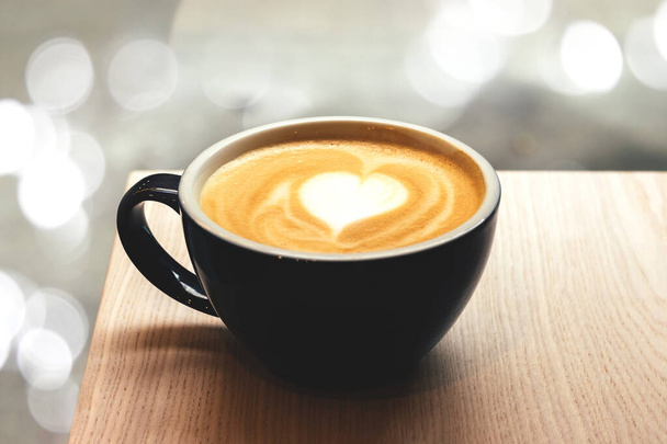 Black cup of coffee with heart shape latte art foam on wooden table in cafe with blurred lights bokeh - Photo, Image