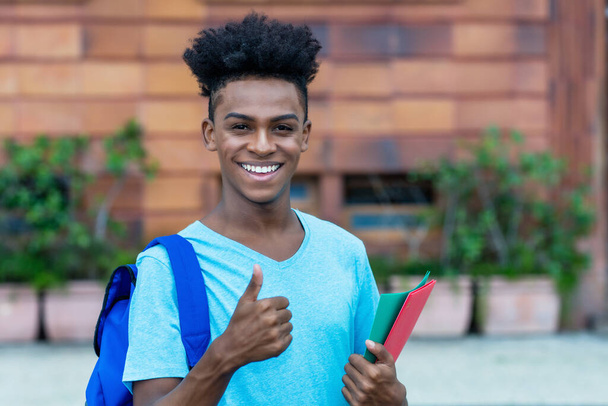 Successful afro american male student with backpack and paperwork showing thumb up outdoor in city in summer - Foto, immagini