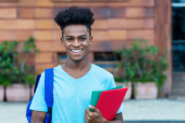 Happy laughing afro american male student with backpack and paperwork outdoor in city in summer - Zdjęcie, obraz