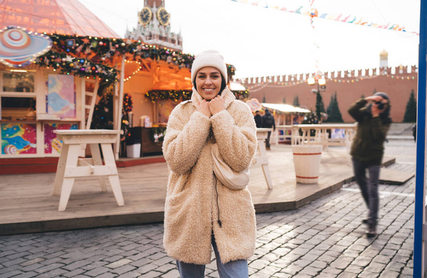 Positive woman in warm coat and hat walking around city during winter holidays and enjoying Christmas decoration and lights around - Fotó, kép