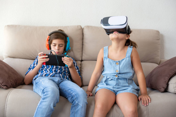 Genç bir çocuk ve küçük bir kız evdeki kanepede farklı dijital teknoloji cihazları kullanıyorlar.. - Fotoğraf, Görsel