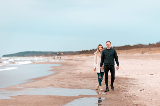 Attractive young couple walking along the shore of a sandy beach, on a spring romantic holiday, outdoors. Travel tourists lifestyle. Couple enjoying loving time together.Copy space. - Foto, Imagen