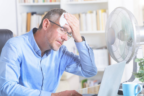 L'uomo soffre di calore mentre lavora in ufficio e cerca di rinfrescarsi dal ventilatore - Foto, immagini