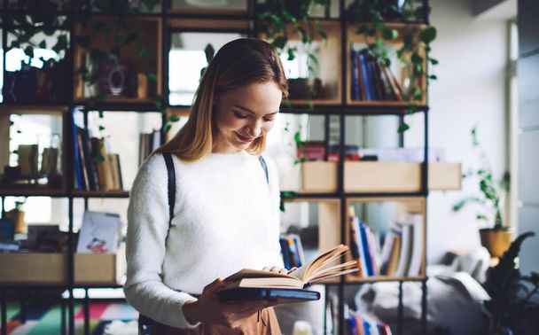 Young female with backpack and in white sweater standing in bright library near bookshelf and reading interesting literature in daytime - Foto, Imagen