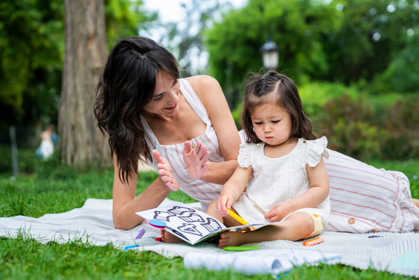 Cheerful young woman with dark hair clapping hands and looking at book with cute little daughter while lying on blanket in park in summer - 写真・画像