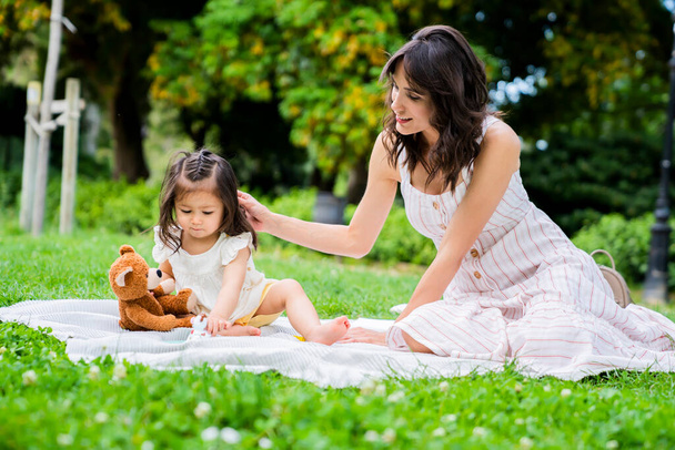 Dark haired young cheerful female in white dress having picnic and patting on head adorable baby girl with teddy bear - Fotó, kép