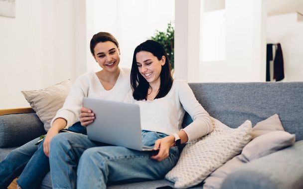 Smiling women in similar outfits using laptop for watching funny videos online while sitting on sofa and spending time together - Foto, Imagem