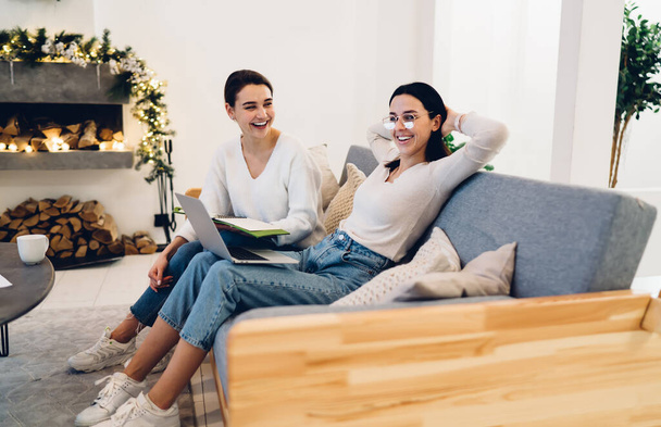 Cheerful female friends in similar clothes sitting on sofa with laptop and notebook while laughing and talking spending time together - Foto, Imagem