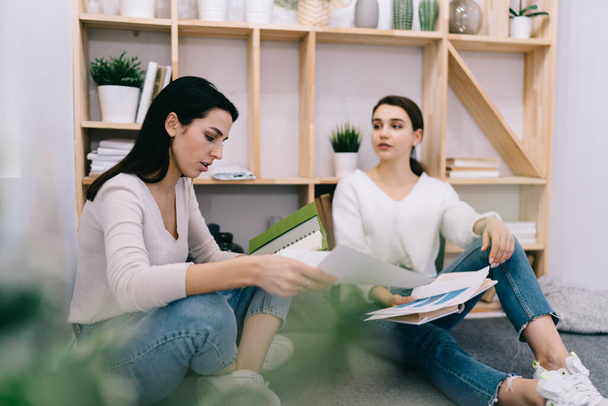 Concentrated female employee looking through documents while discussing work problems to colleague sitting on floor in apartment during informal meeting - 写真・画像