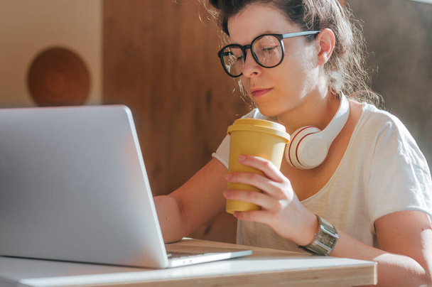 Freelancer feminina usando fones de ouvido trabalhando no laptop e bebendo café. Conceito de geração Z. - Foto, Imagem