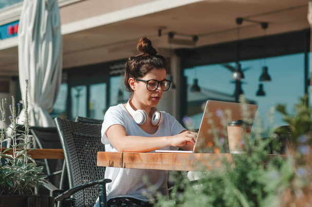 Freelancer feminino usando fones de ouvido trabalhando no laptop no café ao ar livre - Foto, Imagem
