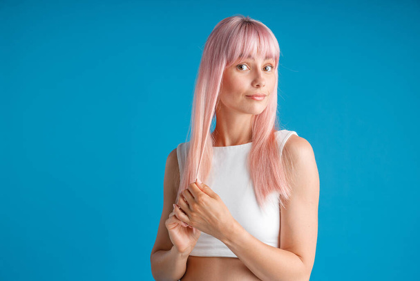 Mladá žena dotýká její hladké přírodní dlouhé růžové barvené vlasy a vypadá překvapeně, pózování izolované přes modré studio pozadí - Fotografie, Obrázek