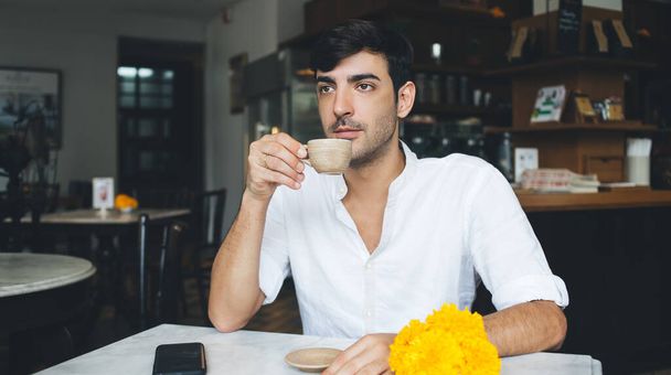 Elegante macho sério em roupas casuais com smartphone olhando para longe e pensando enquanto sentado à mesa tendo bebida quente aromática no café contemporâneo - Foto, Imagem