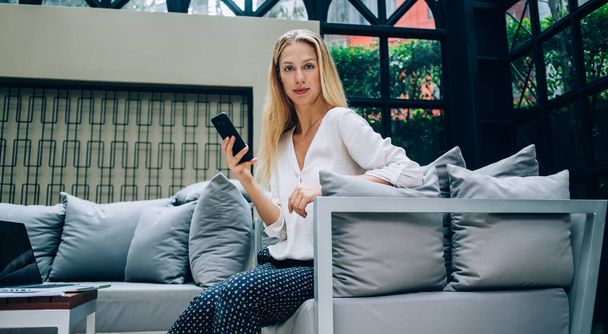 Pensive female entrepreneur in formal clothes browsing smartphone and relaxing in cozy armchair while working remotely and looking at camera - 写真・画像