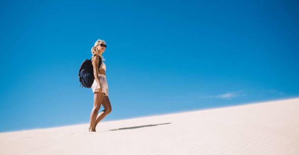 Caucasian female explorer with touristic backpack discovering wild nature sandy environment during summer vacations for recreate, young wanderer standing at Sahara horizon with dunes landscape - Photo, image