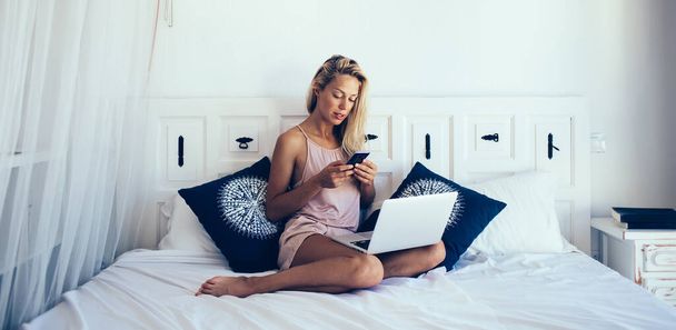 Smart blond haired female freelancer in pajama sitting on bed while surfing on mobile phone and laptop on sunny morning in Barcelona - Фото, изображение