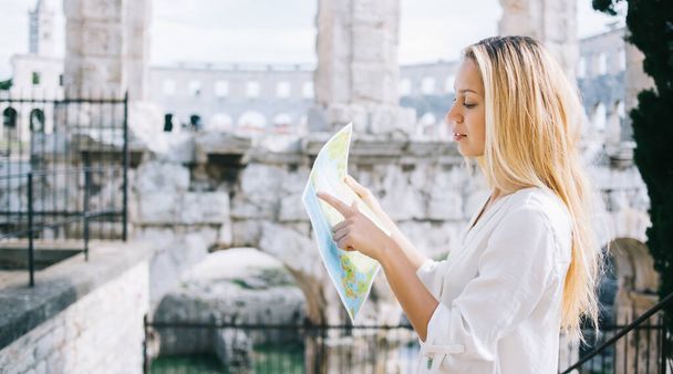 Side view of serious adult woman in casual clothes focusing and pointing finger to paper map while standing against blurred exterior of ancient Coliseum and developing new route in Rome - Foto, Bild