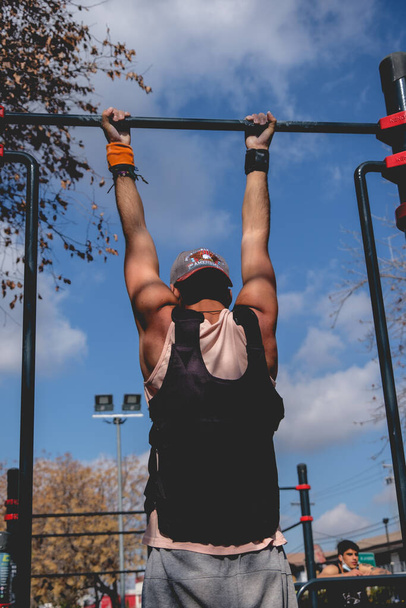 Fit and handsome athlete guy doing a calisthenic exercise routine (performs a pull-up) with a weight vest in an street workout park in a sunny day of summer - Photo, Image