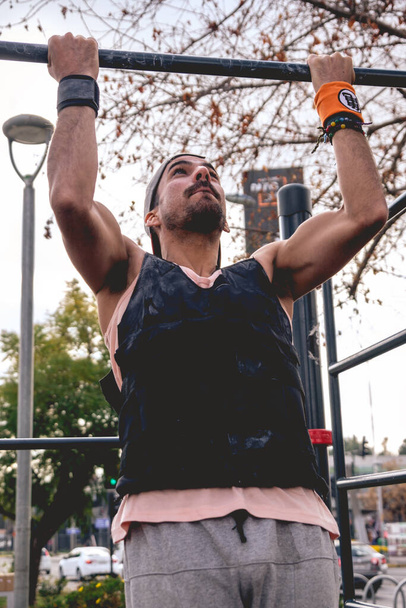 Fit a hezký atlet chlap dělá kalisthenní cvičení rutina (provádí pull-up) s vestou v pouličním parku cvičení v slunečný letní den - Fotografie, Obrázek