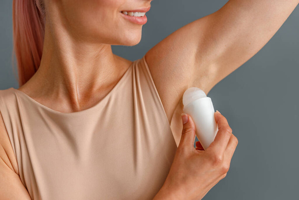 Close up shot of beautiful young woman using antiperspirant and smiling isolated over gray studio background - Foto, afbeelding