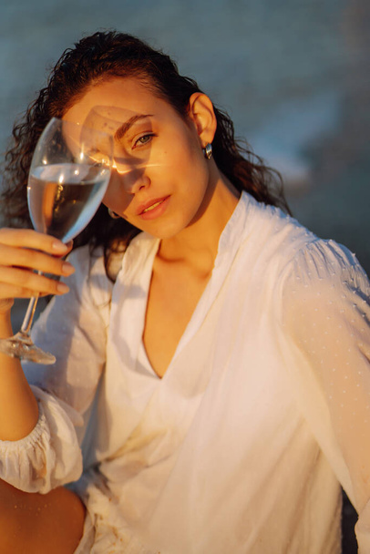Beautiful  model in elegant white dress at sunset with glass of wine. The girl looks at the magical sunrise. Summer time. Travel, weekend, relax and lifestyle concept. - Foto, imagen