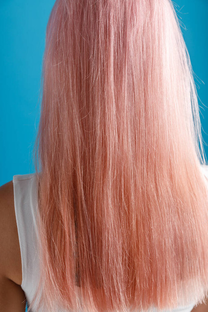 Close up shot of beautiful sleek natural long pink color dyed hair of young woman standing isolated over blue studio background - Фото, изображение