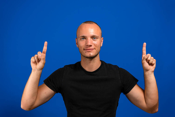 Young caucasian man pointing up with his index fingers while smiling, he is dedicating a good deed to someone who passed away. Isolated on blue studio background. Nostalgia concept - 写真・画像