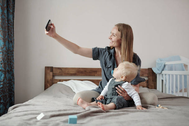 Szczęśliwa młoda matka wspiera plecy syna podczas robienia selfie z nim w łóżku - Zdjęcie, obraz