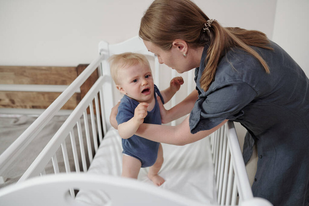 Young mother with ponytail taking frightened baby in arms from crib - Valokuva, kuva