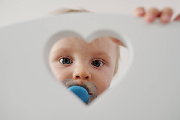 Portrait of cute baby with blue pacifier looking through heart-shaped hole of bed - Foto, Imagen