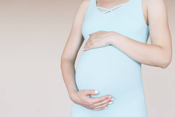 Μια έγκυος γυναίκα αγκαλιάζει το στομάχι της με μπλε ρούχα σε μπεζ φόντο. κοντινό πλάνο - Φωτογραφία, εικόνα